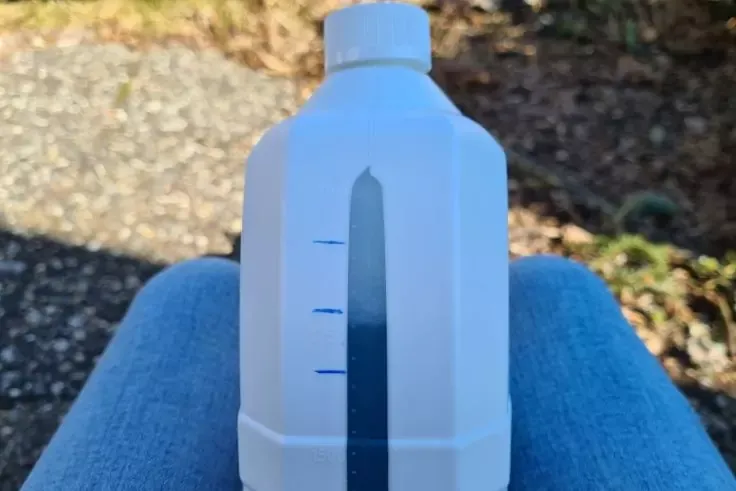 Bottiglia di plastica bianca con tappo a vite e striscia verticale trasparente graduata con segni blu, appoggiata su jeans blu; ghiaia e erba sullo sfondo