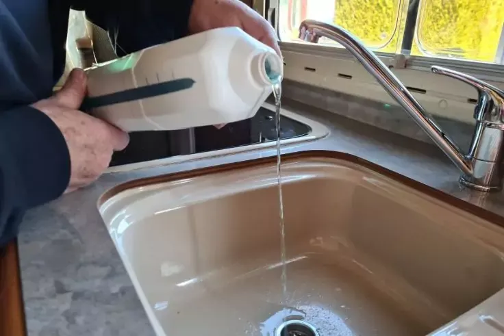 Mani tengono una bottiglia di plastica bianca, acqua scorre in un lavello beige sotto un rubinetto