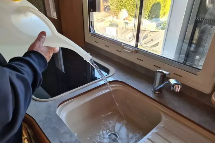 Mano versa acqua da un annaffiatoio bianco in un lavello della cucina sotto una finestra