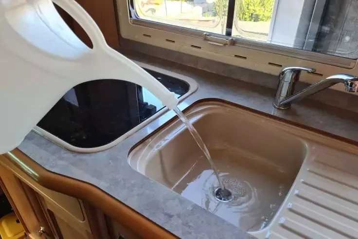 Annaffiatoio in plastica bianco versa acqua in un lavabo rettangolare beige; scarico e getto d'acqua visibili; rubinetto cromato, piano scolapiatti rigato, secondo lavabo a sinistra, finestra sullo sfondo