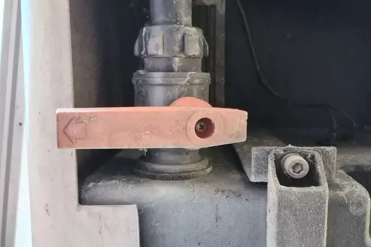 Primo piano di una leva color rame con una freccia su una valvola metallica, montata su una giunzione di tubo grigia, con una staffa metallica impolverata e un cavo nero nelle vicinanze