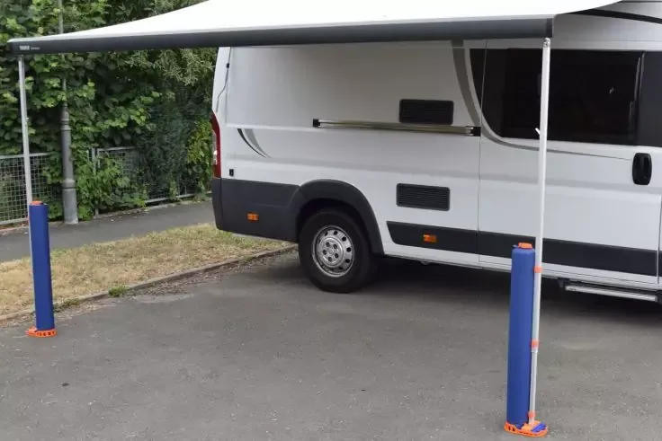 Camper bianco visto di lato con tenda esterna aperta sostenuta da due pali blu, ruota posteriore visibile, parcheggiato su asfalto accanto all'erba, recinzione e alberi sullo sfondo
