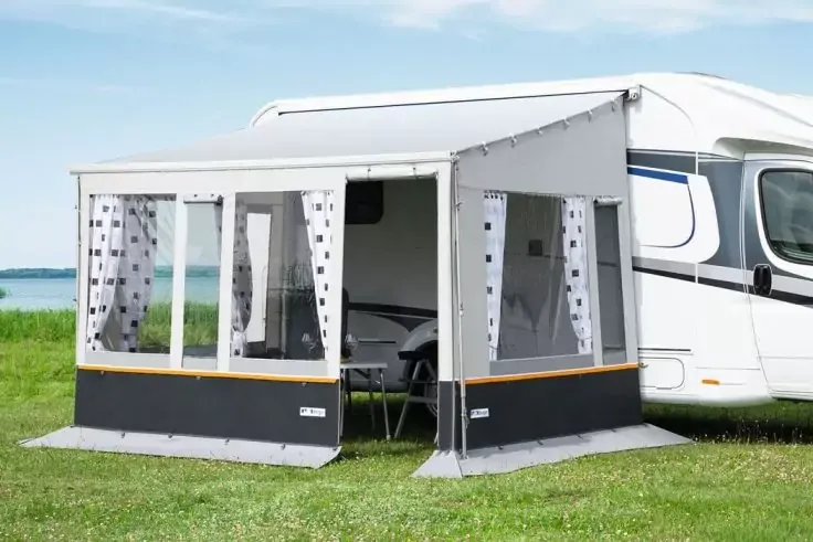 Camper bianco con veranda grigia; due finestre e ingresso aperto; tavolo e due sedie nella veranda; prato e corpo d'acqua sullo sfondo, cielo sereno