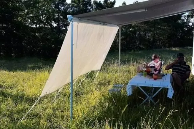 Telo teso a mo' di tenda su pali metallici sopra un tavolo con tovaglia bianca in erba alta; due persone sedute al tavolo; alberi sullo sfondo