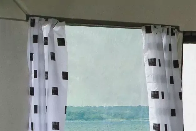 Telaio della finestra con tende bianche a quadrati neri; nella finestra mare e cielo all'orizzonte