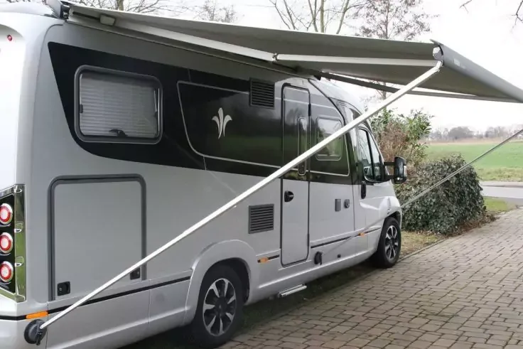 Camper bianco-grigio con tendalino aperto, parcheggiato su una superficie lastricata; cespugli a destra, campo e alberi sullo sfondo
