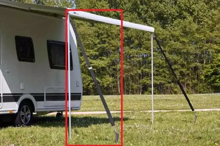 Caravan con tenda estesa e due pali inclinati, erba in primo piano, alberi sullo sfondo