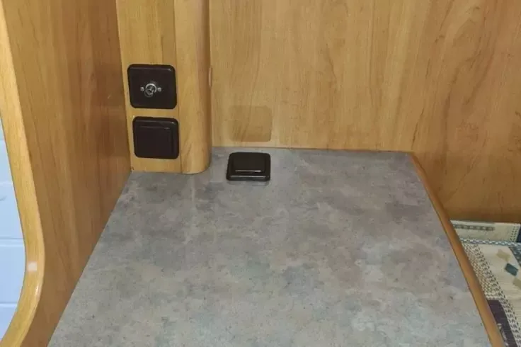 Piano grigio davanti a pannelli di legno; a sinistra due prese nere su un montante in legno; sul piano un piccolo dispositivo rettangolare nero; a destra il bordo di un panno a motivo