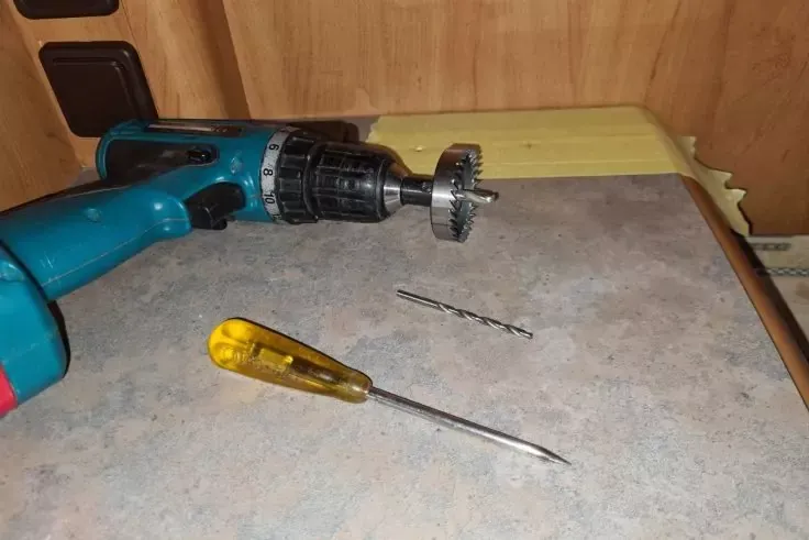 Su un piano di lavoro un trapano elettrico con una sega a tazza montata, una punta da trapano e un cacciavite con impugnatura gialla davanti a un rivestimento in legno