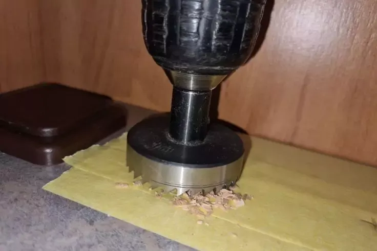 Testa di trapano nera con lama a tazza rotonda su carta gialla su una superficie di lavoro grigia; trucioli di legno sotto l'accessorio metallico, parete in legno sullo sfondo, coperchio di plastica marrone a sinistra