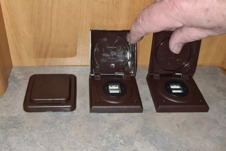 Tre custodie quadrate marroni in plastica su un piano di lavoro; due aperte, ognuna con una sede rotonda e un piccolo dispositivo nero con display rettangolare; custodia sinistra chiusa; una mano indica la custodia aperta a destra