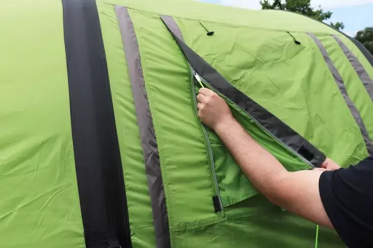 Braccio e mano aprono la cerniera di una patta su una tenda verde con strisce verticali scure