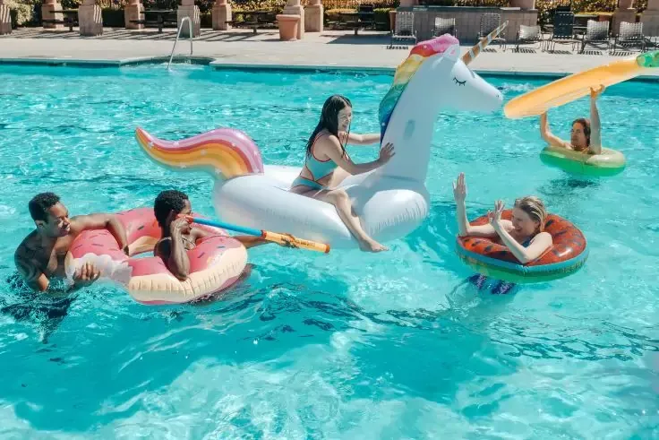 Cinque persone su gonfiabili in una piscina: una su un grande unicorno bianco, due su una ciambella rosa, una su una ciambella rossa, una su una tavola gialla; sdraio e ombrelloni a bordo piscina