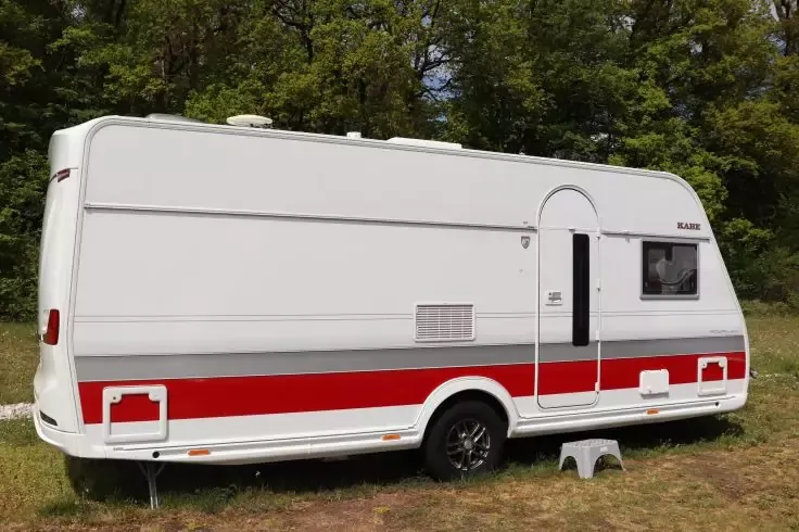 Caravan bianco con striscia rossa, porta aperta, finestra, ruota e piccolo gradino, su erba davanti ad alberi
