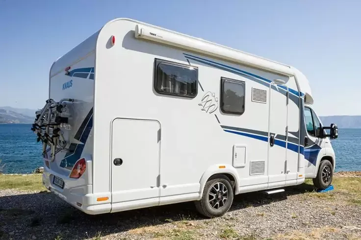 Camper bianco con strisce blu, finestrini laterali, cassone della tenda e portabici posteriore su ghiaia vicino al mare; acqua e montagne in lontananza, cielo sereno