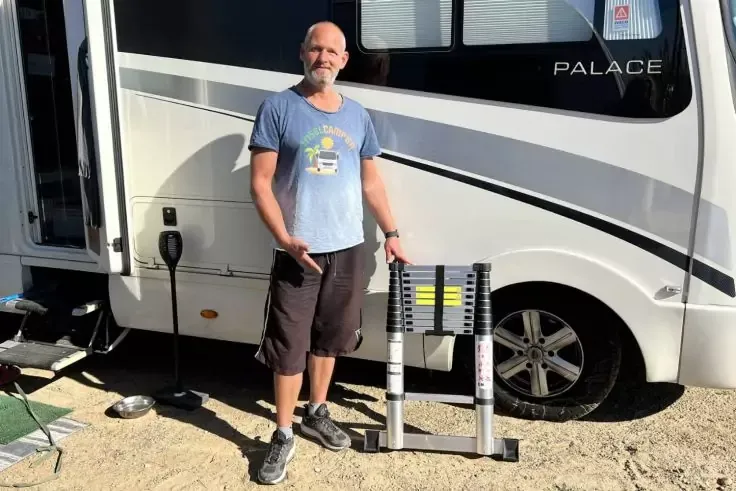 Un uomo &egrave; accanto a un camper bianco e tiene una scala telescopica piegata; ruota, porta aperta, cavo e una ciotola metallica su terreno sabbioso