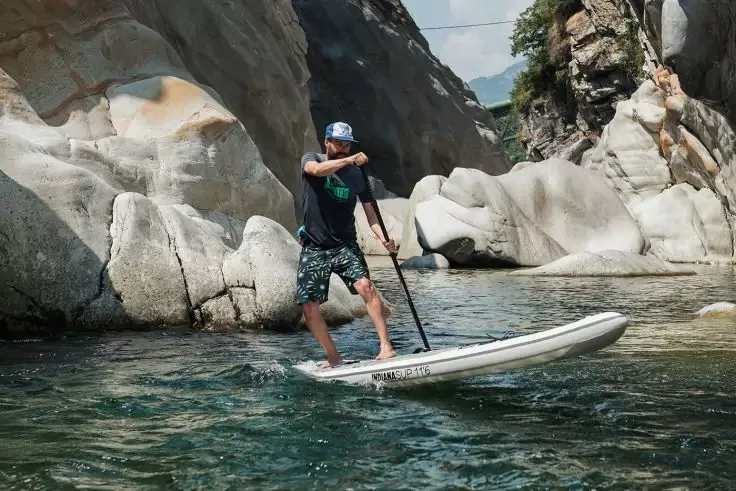 Persona in piedi su una tavola da stand-up paddle in acqua di fiume limpida tra formazioni rocciose, indossa casco, maglietta e pantaloncini, tiene una pagaia