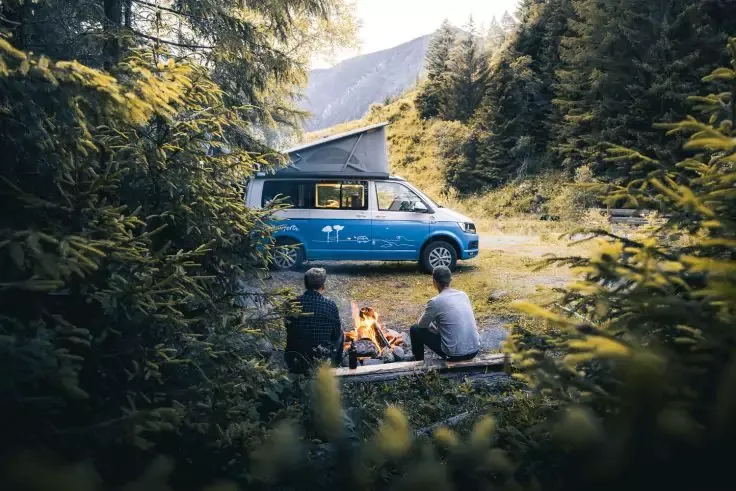 Furgone camper blu con tetto sollevato su un prato; in primo piano due persone sedute su tronchi vicino a un fal&ograve;; conifere e una montagna sullo sfondo