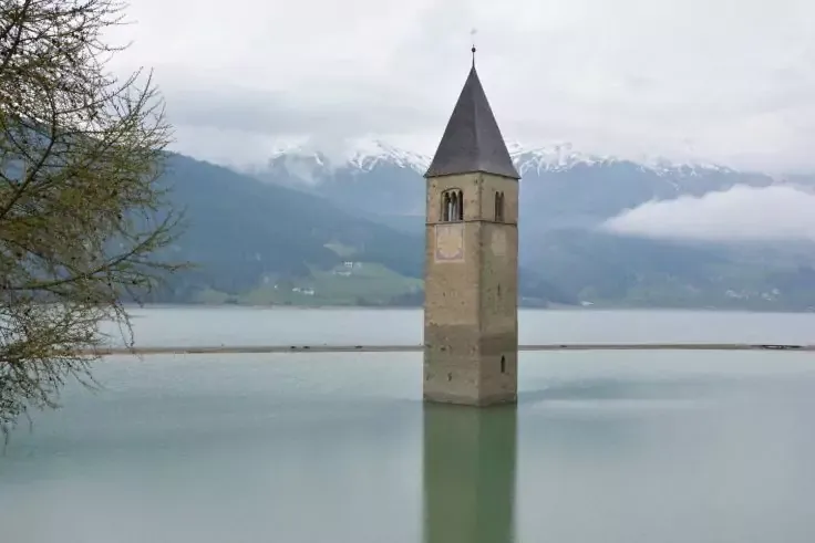 Torre di pietra con tetto a punta nel lago calmo, riflesso nell'acqua, montagne innevate e cielo nuvoloso sullo sfondo, ramo d'albero a sinistra