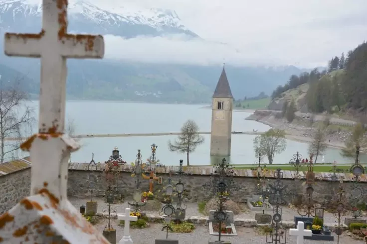 In primo piano una croce invecchiata a sinistra; cimitero con croci e lapidi dietro un muro di pietra; lago con un campanile nell'acqua; riva con alberi; montagne innevate e nebbia sullo sfondo; cielo nuvoloso