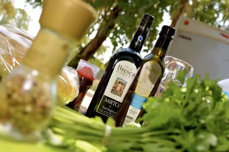 Due bottiglie di vetro scuro con etichette, in primo piano erbe verdi fresche, accanto un bicchiere e un barattolo di vetro, sfondo sfocato con fogliame