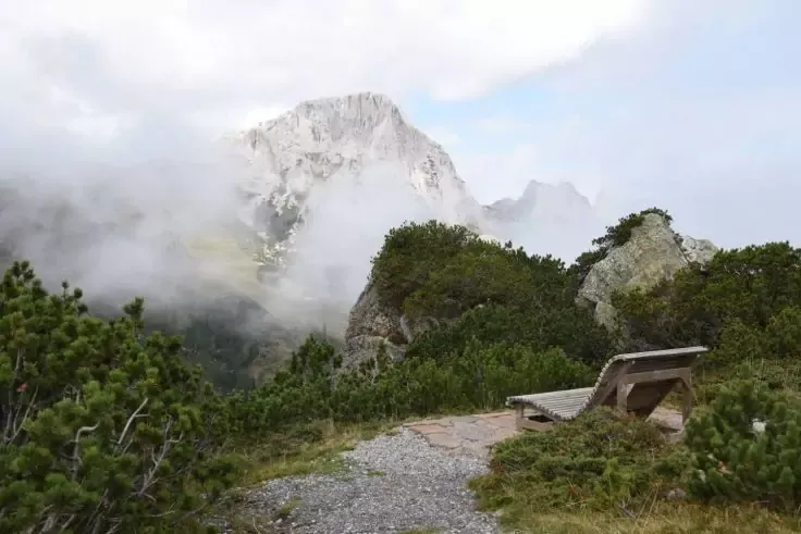 Panchina di legno su un sentiero ghiaioso, arbusti verdi bassi in primo piano, cima rocciosa parzialmente coperta da nebbia e nuvole