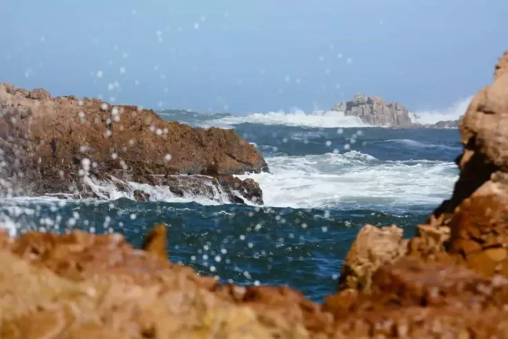 Costa rocciosa in primo piano, onde e spruzzi d'acqua, scogli in lontananza, cielo azzurro