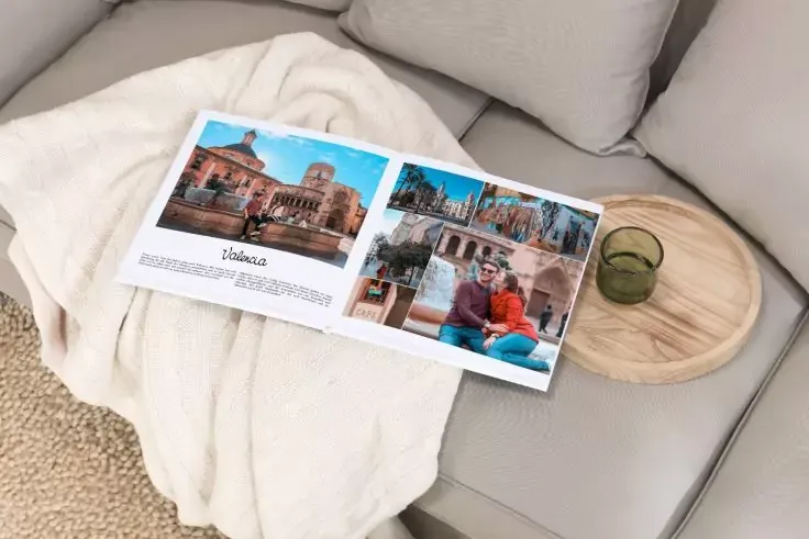 Libro fotografico aperto con immagini di edifici e una persona su un divano chiaro con una coperta chiara, accanto un vassoio tondo di legno con un bicchiere verde