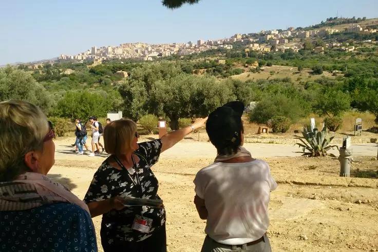 Tre persone viste di spalle/di profilo, una persona indica con il braccio teso; terreno sabbioso con piante di agave e piccola vegetazione; alberi su colline ondulate e case sullo sfondo, cielo azzurro