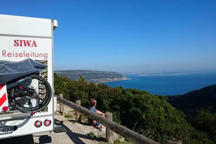 Camper bianco con la scritta 'SIWA Reiseleitung' e una bicicletta sul retro, ringhiera in legno e cespugli in primo piano, una persona seduta vicino alla ringhiera; mare blu, scogliere costiere e cielo sereno sullo sfondo
