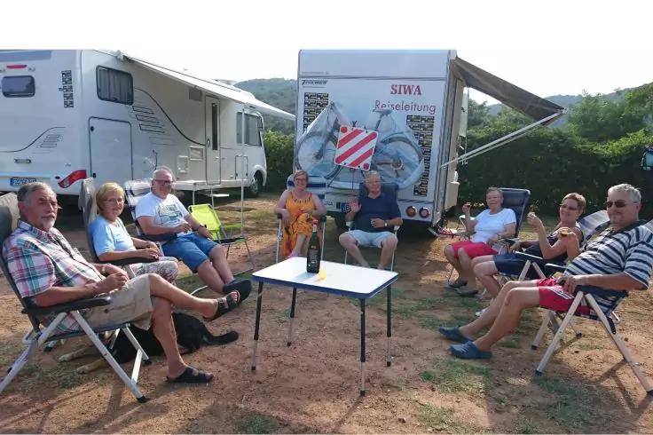 Nove persone sedute a mezzaluna su sedie pieghevoli intorno a un tavolino bianco con bottiglie e bicchieri, davanti a due camper e un rimorchio con tenda estesa su terreno rossastro, sullo sfondo colline e vegetazione