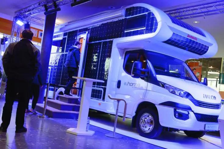 Camper bianco con pannelli solari scuri sul fianco e sul tetto, sportello laterale aperto con apparecchiature visibili, due persone nelle vicinanze, illuminazione d'esposizione blu in una hall