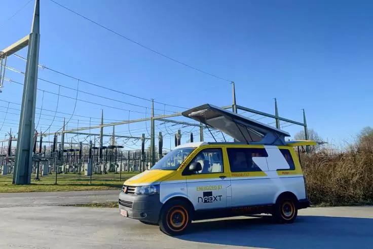 Furgone giallo e bianco con tetto sollevato e logo "next" su una superficie asfaltata davanti a una cabina elettrica con linee elettriche, cielo blu