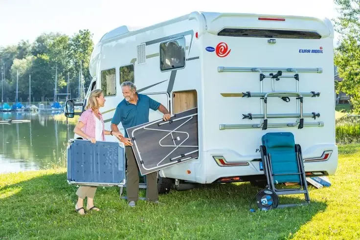 Twee personen tillen een opgevouwen tafel uit het achtervak van een witte camper; daarnaast een opgevouwen campingstoel, gras, meer met boten en bomen