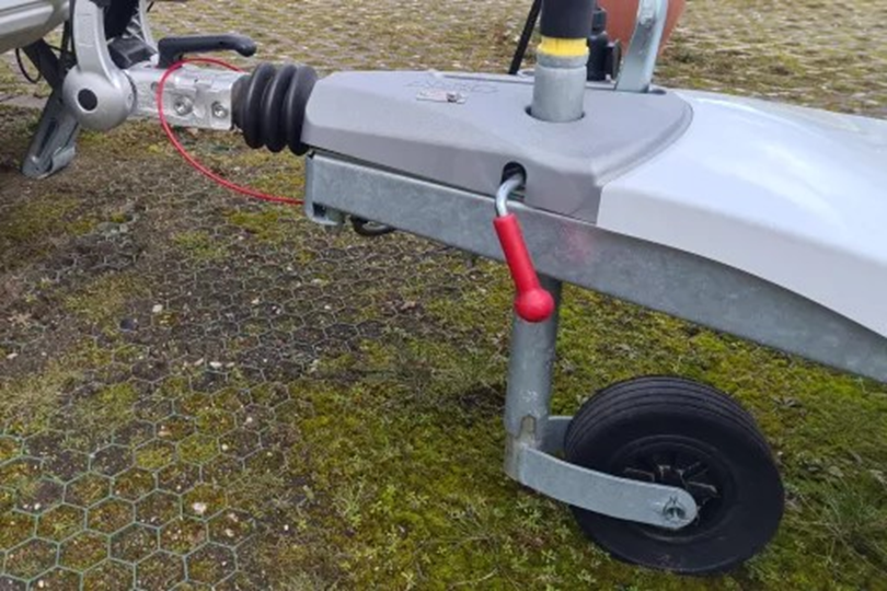 Aanhangerdissel met kogelkoppeling, grijs steunwiel met zwarte band, rood handvat aan een kabel, zeshoekige bestrating met mos