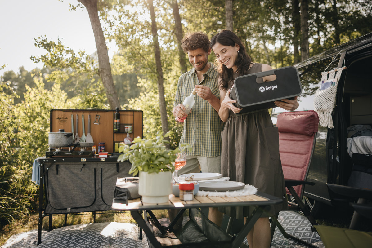Zwei Personen (ein Mann und eine Frau) stehen an einem Campingtisch; auf dem Tisch Teller, Besteck, eine Topfpflanze und Getr&auml;nke; im Hintergrund ein offenes Fahrzeug mit Campingausr&uuml;stung, ein Klappstuhl und B&auml;ume