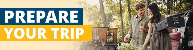 Two people stand beside a car with an open trunk; left a banner reading 'REISE-VORBEREITUNG'; trees in the background