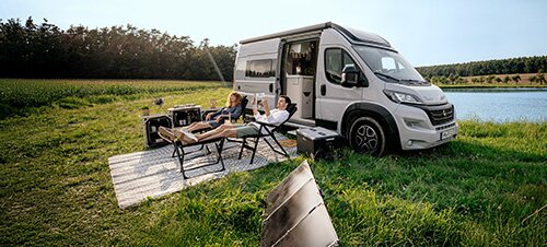 Zwei Personen entspannen auf Liegestühlen neben einem Camper in einer grünen Landschaft am See