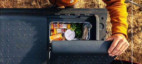 Person mit orangefarbenem Pullover öffnet schwarzen Kunststoffbehälter mit drei orangen Getränkekartons, einem weißen Becher mit Deckel, Salatblättern und einem verpackten Sandwich