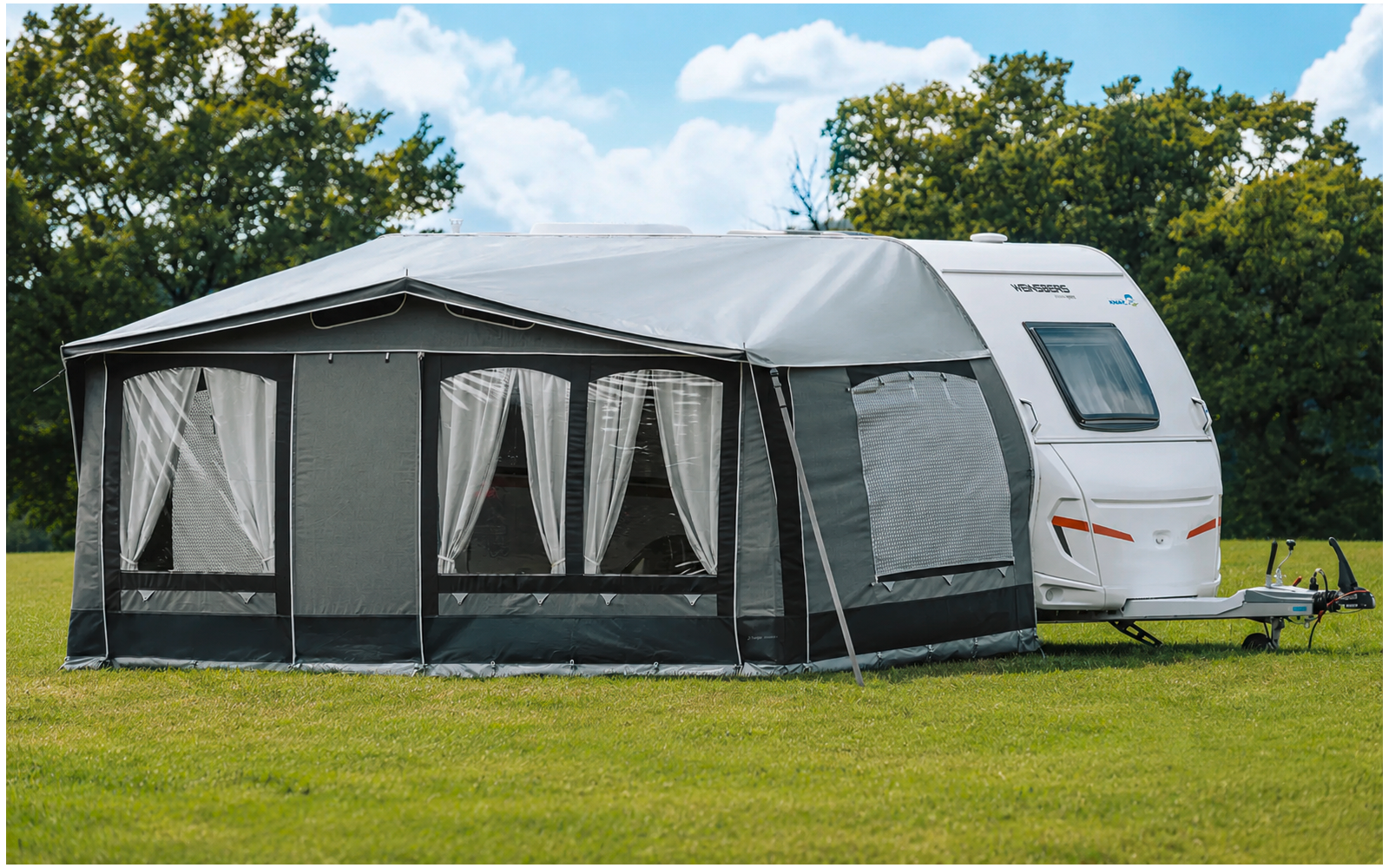 White caravan with a gray awning attached on green grass; awning has three windows with white curtains; trees and blue sky in the background