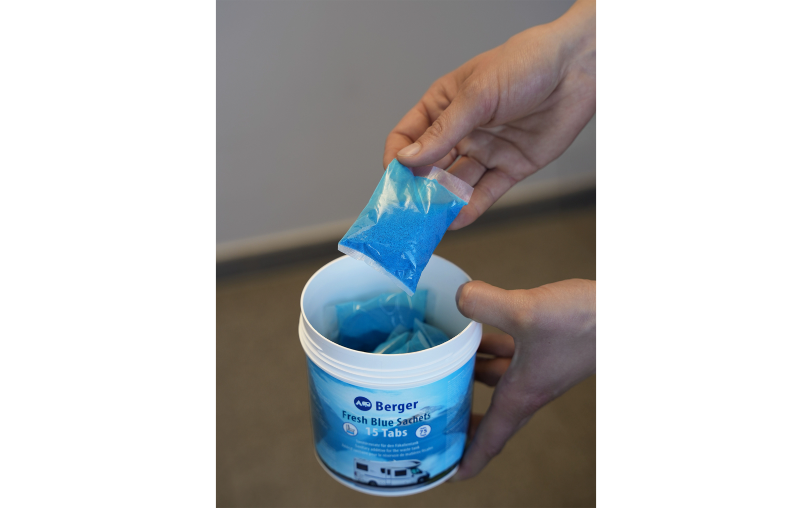 Twee handen houden boven een open witte bus met blauw etiket een klein transparant zakje met blauwe korrels; in de bus liggen meer vergelijkbare zakjes