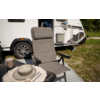 Grijze inklapbare campingstoel met armleuningen vóór de zijkant van een witte caravan; kleine zwarte tafel met een beige bord, kom en mok op een lichte doek, geprint vloermat, gras en bomen op de achtergrond