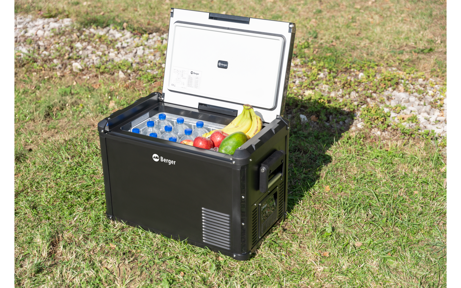 Glacière portable noire au couvercle blanc ouvert sur l'herbe; à l'intérieur bouteilles d'eau, bananes, pommes et un concombre