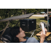 Personne allongée sur une chaise longue tenant un smartphone; grand appareil noir sur un support, anneau lumineux et table pliante dans un parc