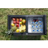 Glacière noire ouverte sur l'herbe; côté gauche avec pommes, citrons, bananes, un ananas et un fruit vert; côté droit avec de la glace, plusieurs bouteilles en plastique avec bouchons bleus et deux bouteilles foncées avec bouchons rouges