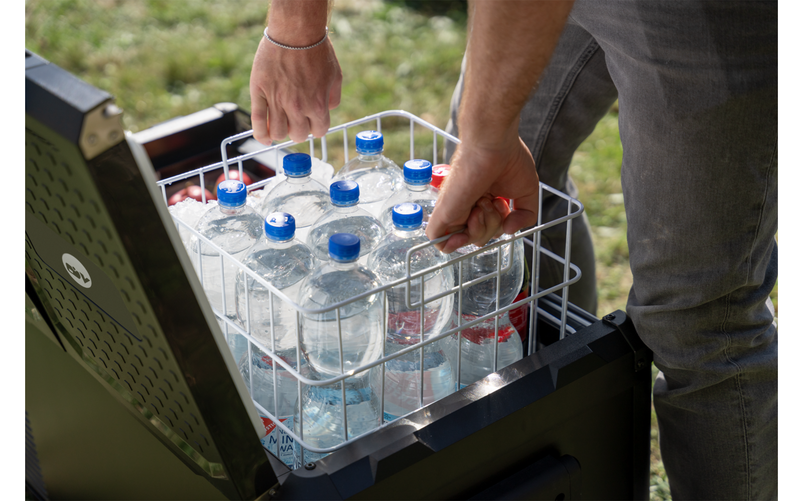 Mains d'une personne saisissant une bouteille en plastique dans un panier métallique contenant plusieurs bouteilles d'eau à l'intérieur d'une glacière ouverte, herbe en arrière-plan