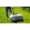 Lower body of a person in dark jeans and black shoes pulling a gray-white plastic wheeled box with a telescopic handle and the label "Berger" across green grass