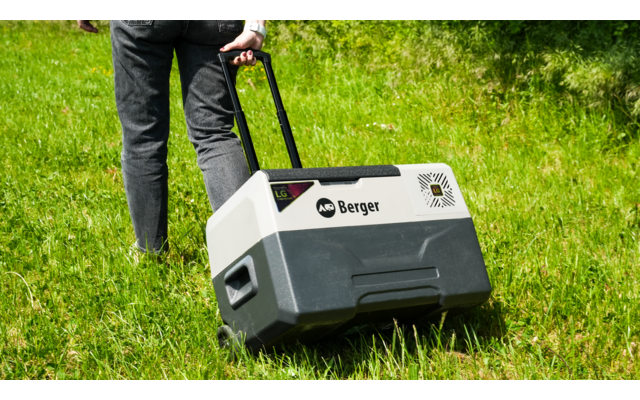Lower body of a person in dark jeans and black shoes pulling a gray-white plastic wheeled box with a telescopic handle and the label "Berger" across green grass