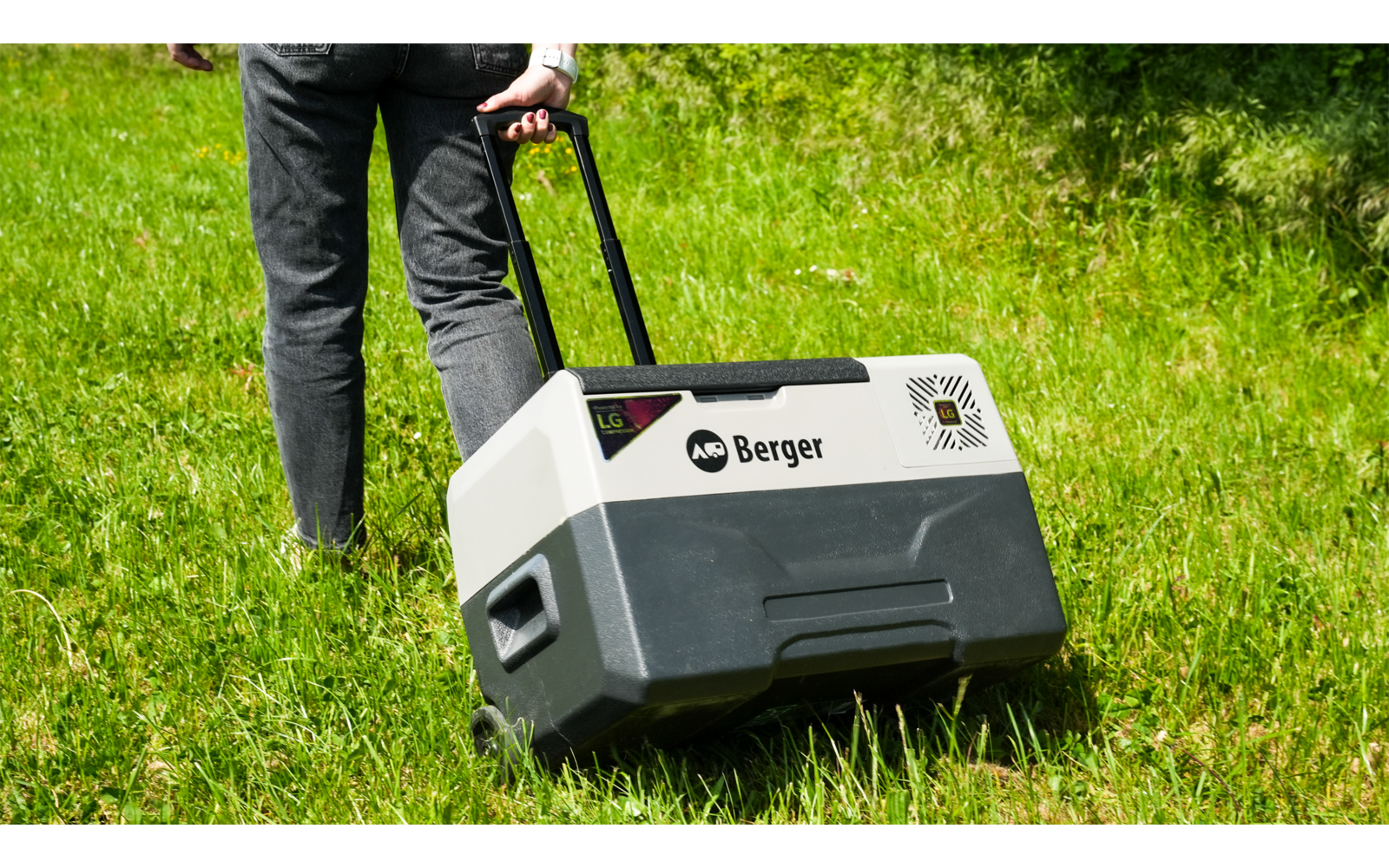 Lower body of a person in dark jeans and black shoes pulling a gray-white plastic wheeled box with a telescopic handle and the label "Berger" across green grass