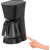 Cafetera de goteo negra con jarra de vidrio; una mano derecha pulsa un botón de encendido rojo